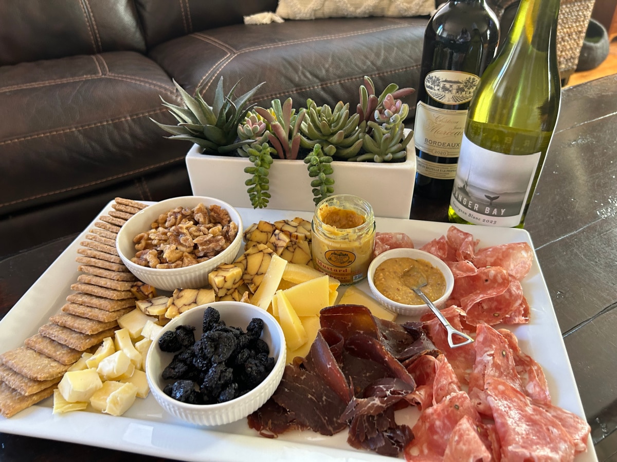 A white platter of meats and cheeses arranged with dip and two bottles of wine
